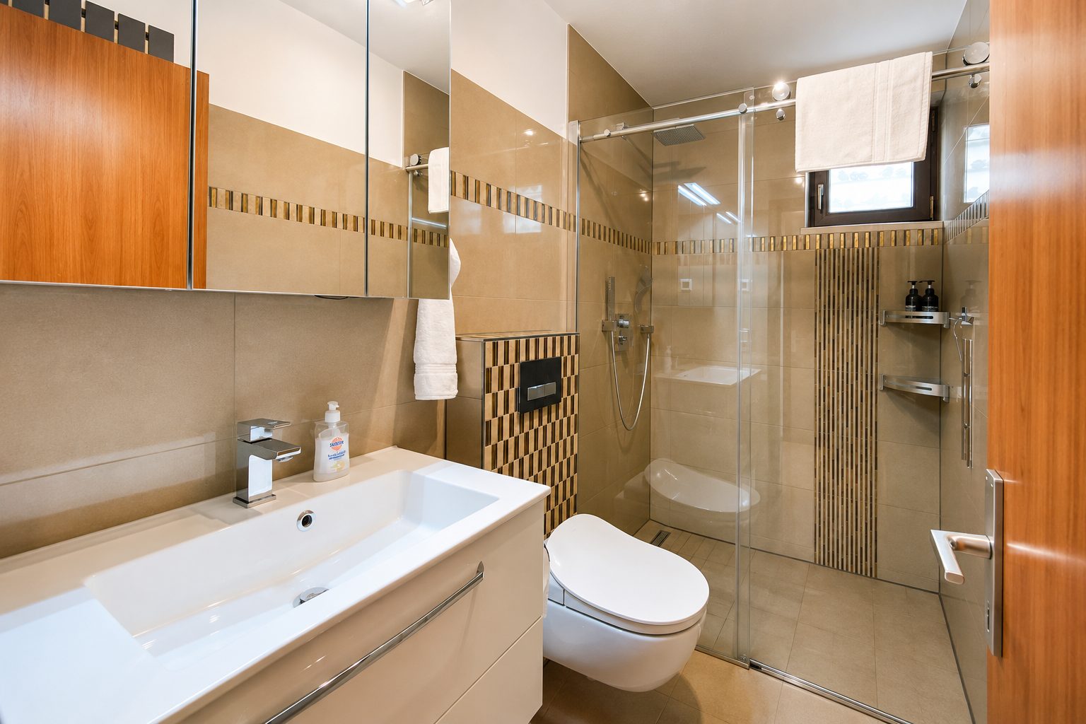 Badkamer in hout- en beigetinten met wastafel, hangtoilet en grote glazen douche met mozaïekrand