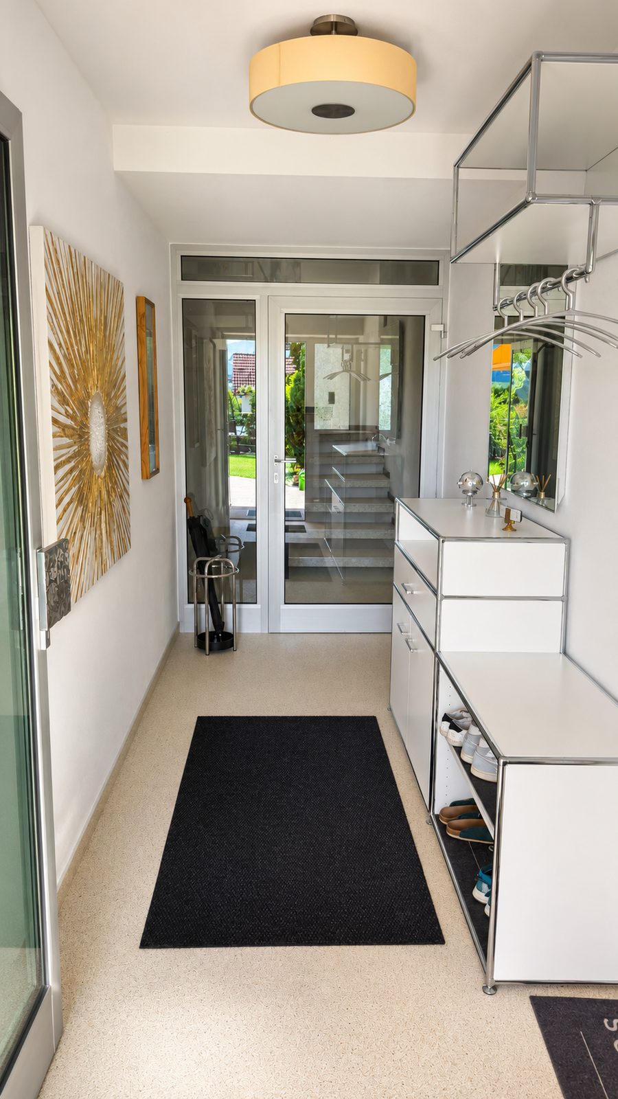 Entreehal met witte modulaire dressoirkast, garderobe met hangers, designlamp, wanddecoratie met zon en glazen deur met uitzicht op het trappenhuis