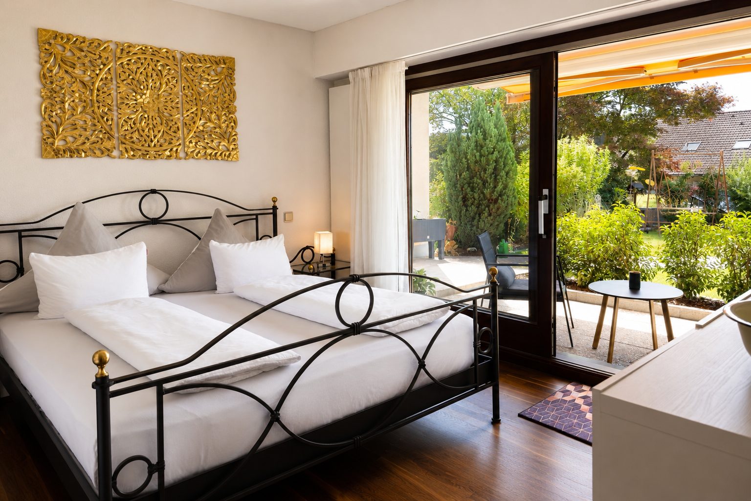 Slaapkamer met zwart metalen tweepersoonsbed, gouden driedelig wandornament, nachtkastlamp en grote glazen schuifdeur met uitzicht op de tuin