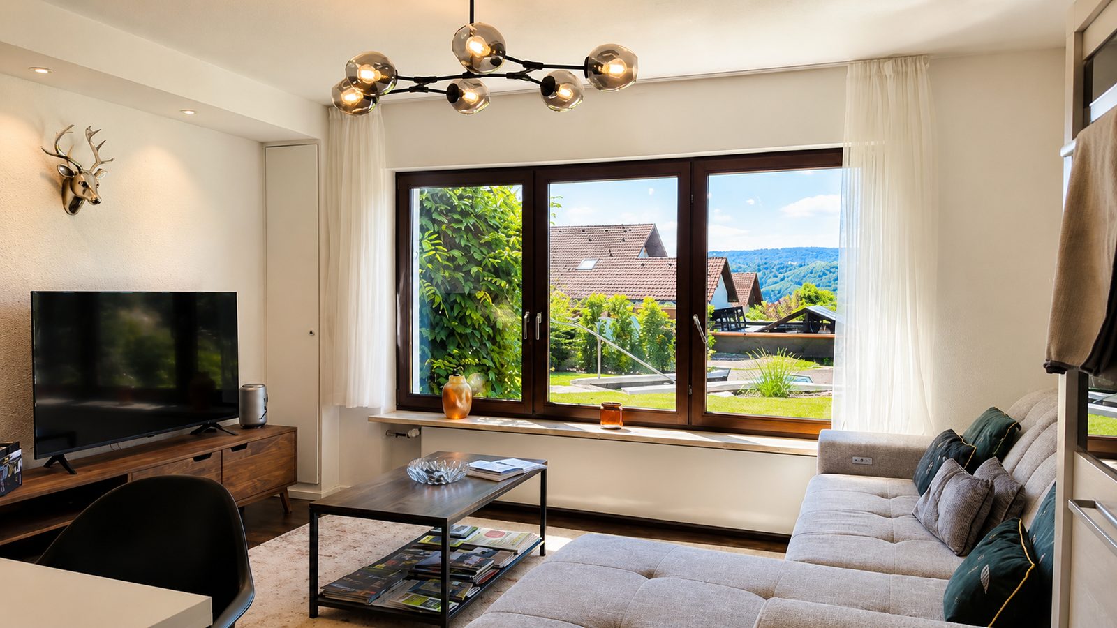 Woonkamer met tv-meubel, bank en groot panoramaraam met uitzicht op huizen en het heuvellandschap