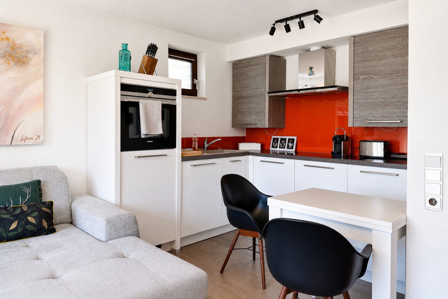 Open woon- en keukengedeelte met bank, kleine eettafel met zwarte kuipstoelen en witte inbouwkeuken met rode glazen achterwand